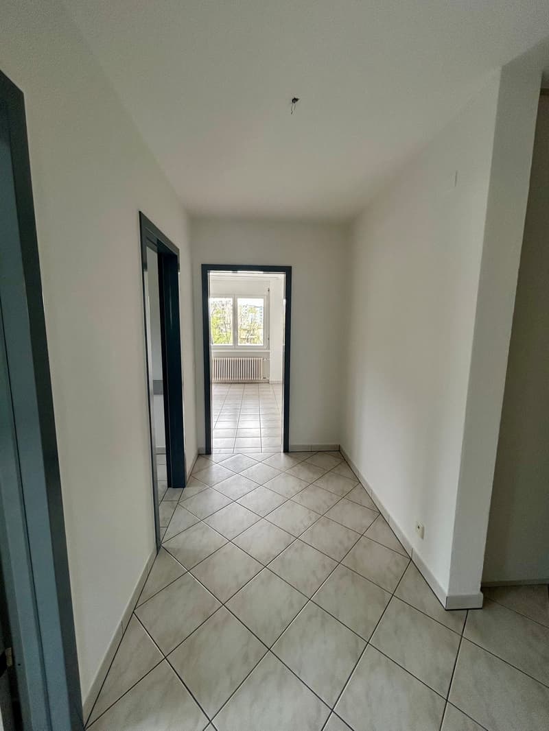 The image shows a hallway with white walls and a tiled floor in a diagonal pattern. There are two doors visible, one on the left and one at the end of the hallway leading to another room.