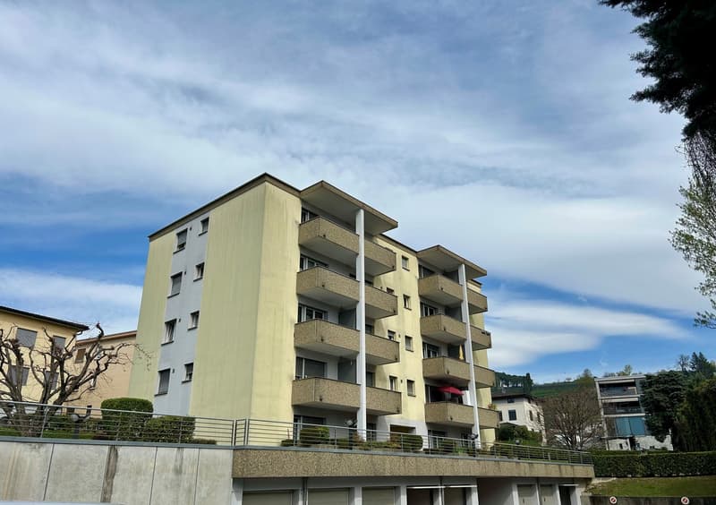 Multi-story apartment building with balconies, beige exterior, and surrounded by trees and greenery.