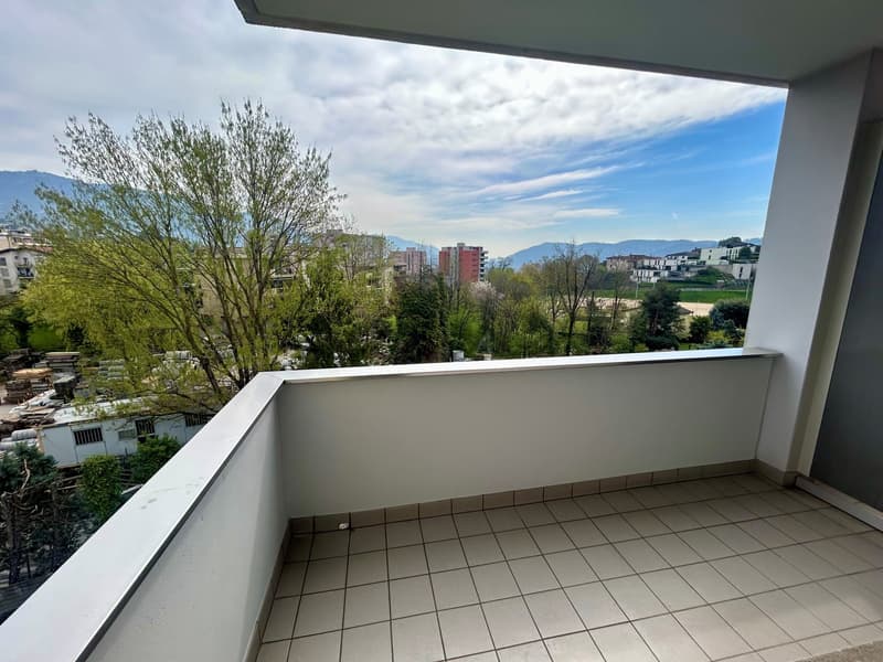 Spacious balcony with a view of the surrounding neighborhood, including trees, buildings, and mountains in the distance. The balcony has a tiled floor and a railing.
