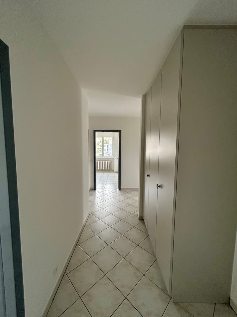 The image shows a hallway with white walls and a tiled floor in a diagonal pattern. There is a closet or cabinet along one wall, and a doorway leading to another room at the end of the hallway.
