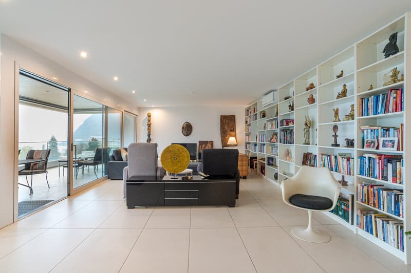 Minimalist living room with white walls, tile floor, large glass doors, couch, center table, bookshelf, armchairs, coffee table, side table, lamp, outdoor dining table and chairs, decorative items, scenic view of mountains and a building