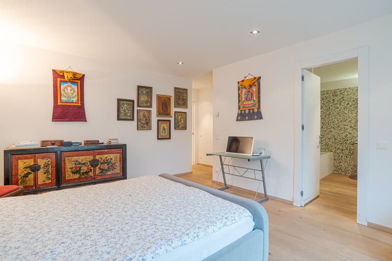 a bed, wooden floor, a desk with a laptop, a cabinet with various items, multiple frames hanging on the wall, an open door to another room