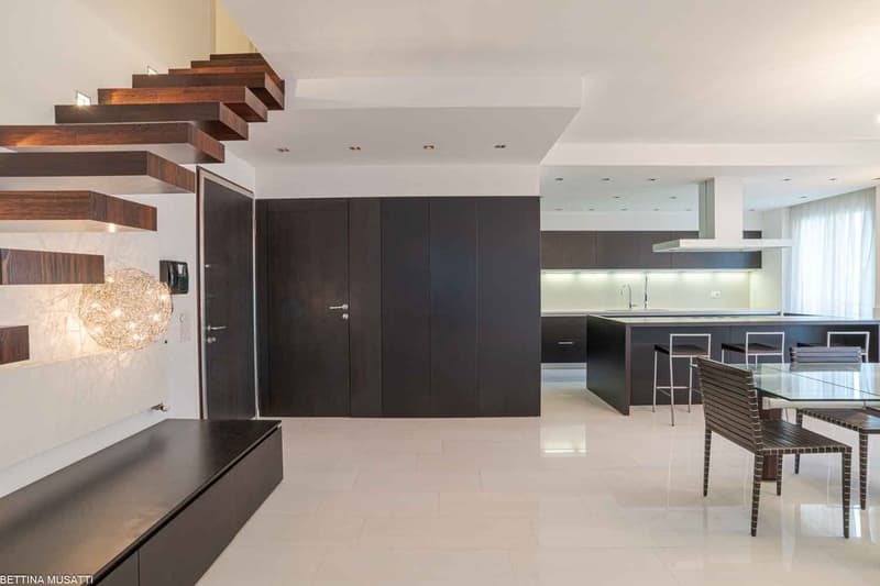 Modern, bright interior with wooden staircase, marble floors, open kitchen, dining area, and minimalist furnishings.
