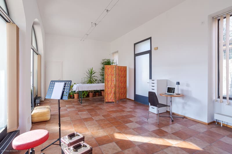 modern interior, white walls, arched windows, terracotta tiles, indoor plants, desk with laptop, chair, music stand, red stool, suitcases