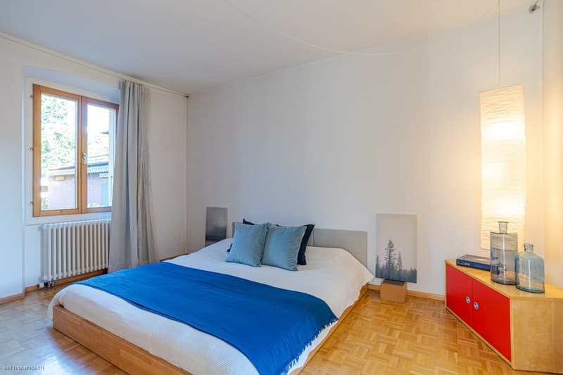 A double bed, white walls, hardwood floor, white radiator, a glass window with curtains, a nightstand with a lamp, a wooden chest, a red cupboard with two doors, and two clear vases on top.