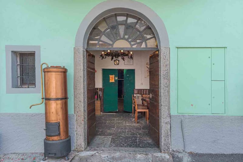 Exterior of a building with green doors and brick walls, featuring a copper pipe next to the entrance and an open doorway with a chandelier.