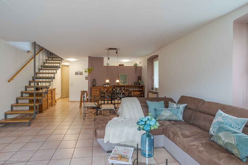 Spacious living room with sofa, wooden staircase, tile floor, wooden dining table, dining chairs, wooden cupboard, kitchen cabinets, flower vase, picture frames, chandeliers, books, and magazine.