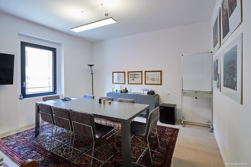 A rectangular conference table with chairs and a rug, a cabinet, a white board, pictures, a TV, and a lamp