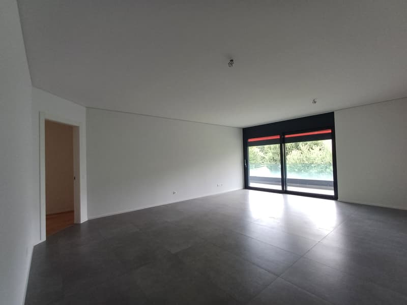 Large open space with white walls, gray tile flooring, and a sliding glass door leading to an outdoor area. The room appears to be empty, with no furniture or decor visible.