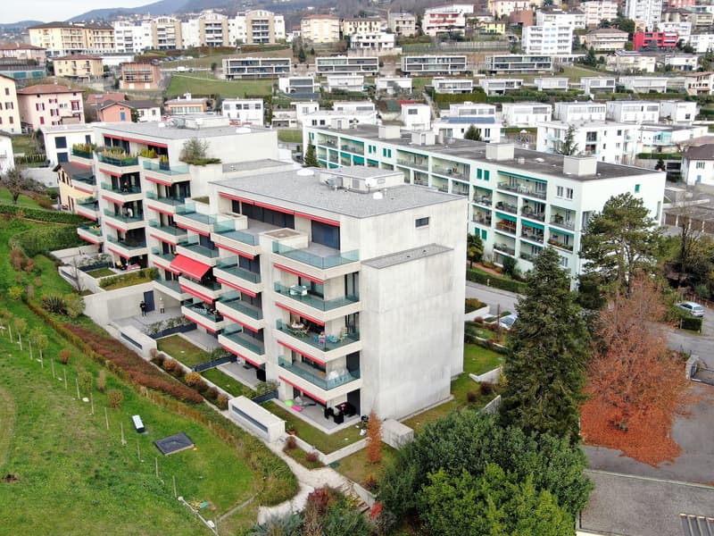 Multi-story apartment building with white exterior and red accents, balconies on each unit, surrounded by greenery and other residential buildings