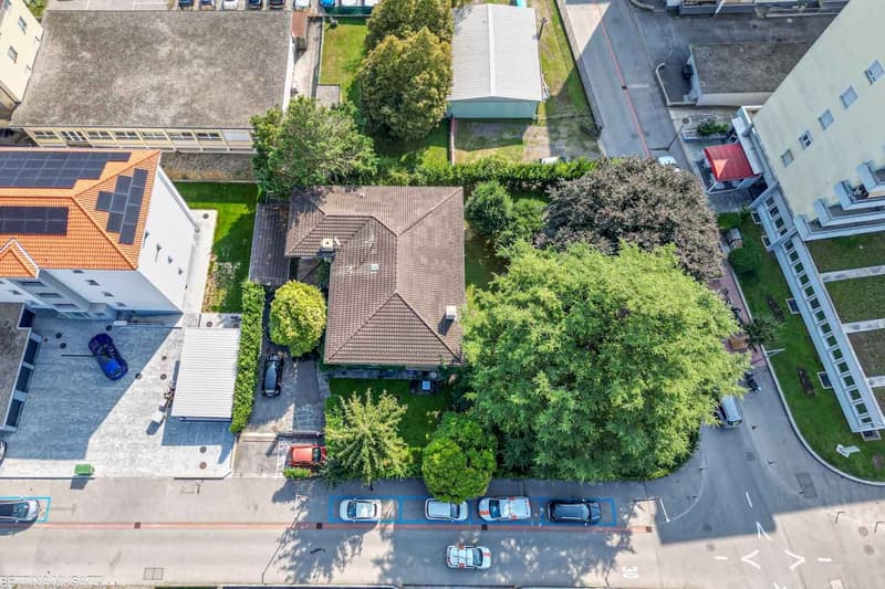 House with brown roof, surrounded by trees and greenery, multiple cars parked