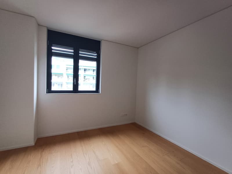 Empty bedroom with white walls, hardwood flooring, and a large window with black frames.