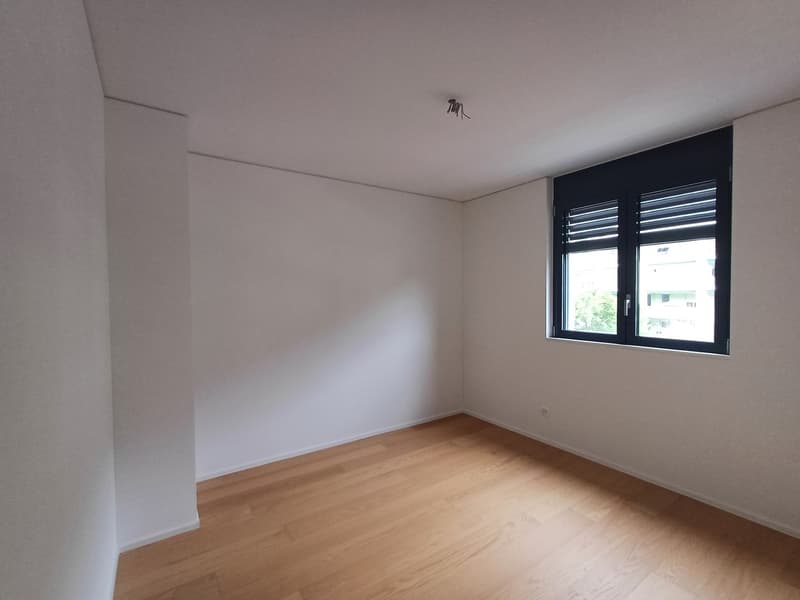 Spacious bedroom with white walls, hardwood flooring, and a large window with black shutters providing natural light