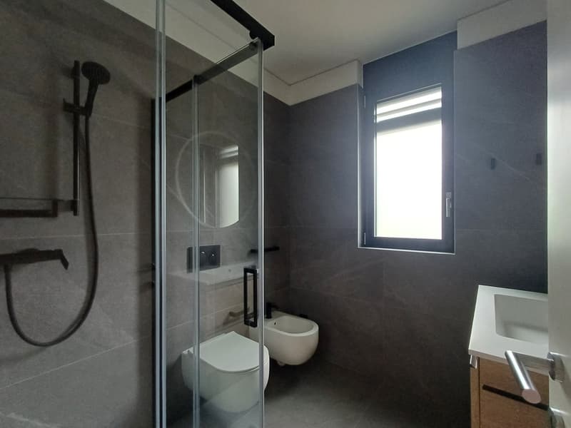 Modern bathroom with glass shower enclosure, white toilet, vanity with sink, and a window providing natural light.
