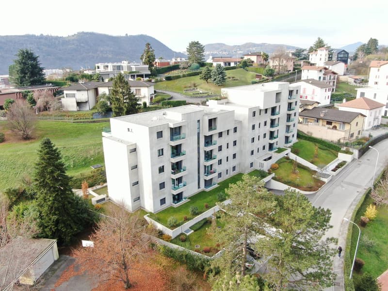 Multi-story apartment building with balconies, surrounded by landscaped gardens and greenery, with mountains in the background