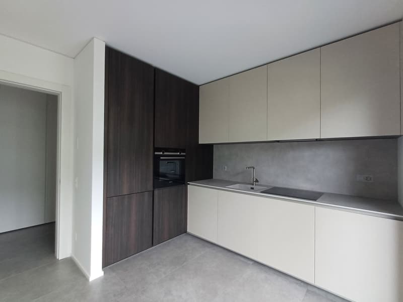 Modern kitchen with white and dark wood cabinets, built-in oven, and a sink with a faucet. The countertop appears to be made of a gray material, possibly concrete or stone. The floor is covered with a light-colored tile or laminate flooring.