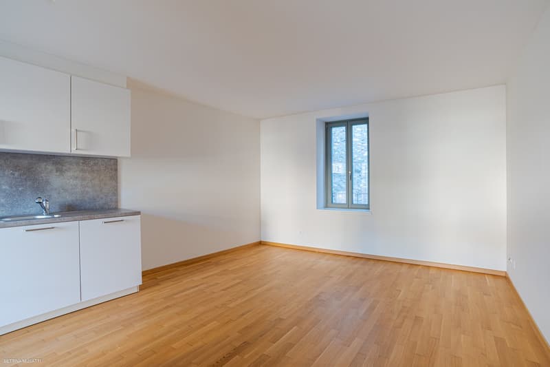 room, hardwood floors, white walls, window, kitchen area with sink and cabinets