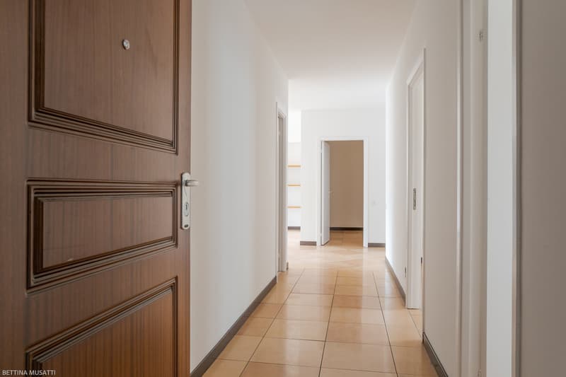 wide hallway, light brown tiles, white walls, wooden door, closed doors, corner room, open entrance