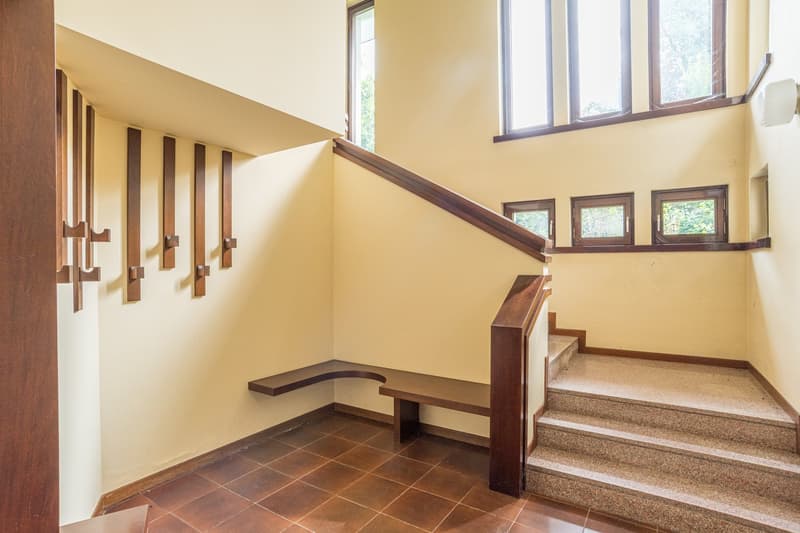 granite stairs, wooden handrail, bench at the bottom of the stairs, beige walls, wooden brackets on the wall, windows on the wall