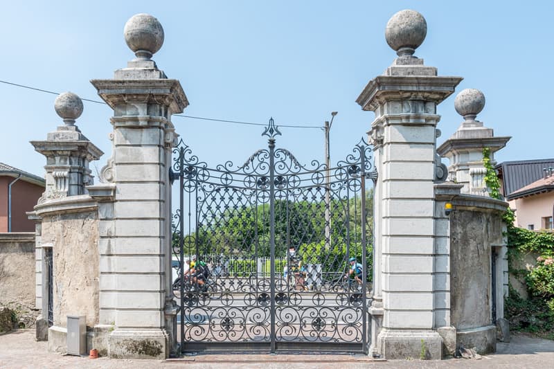 Grand old entrance gate with stone pillars, wrought iron gates, small wall-mounted lamps, bicycle riders at the entrance.