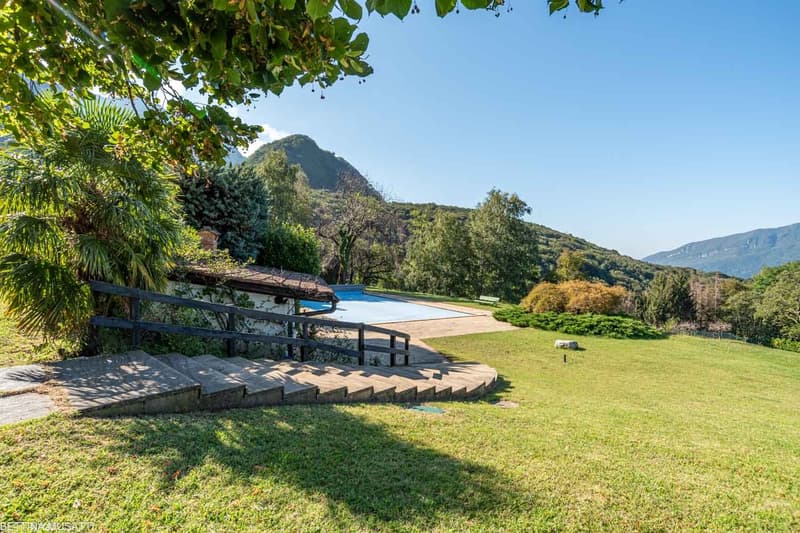 Large open grassy area with a swimming pool, concrete steps leading to the building, surrounded by greenery and distant hills
