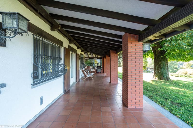 An outdoor area, with a wooden walkway covered by a tiled roof supported by brick pillars. The walkway has arched wooden beams. There are chairs and a griller next to the entrance door, with a glass window on the wall that has an exterior black metal fenc