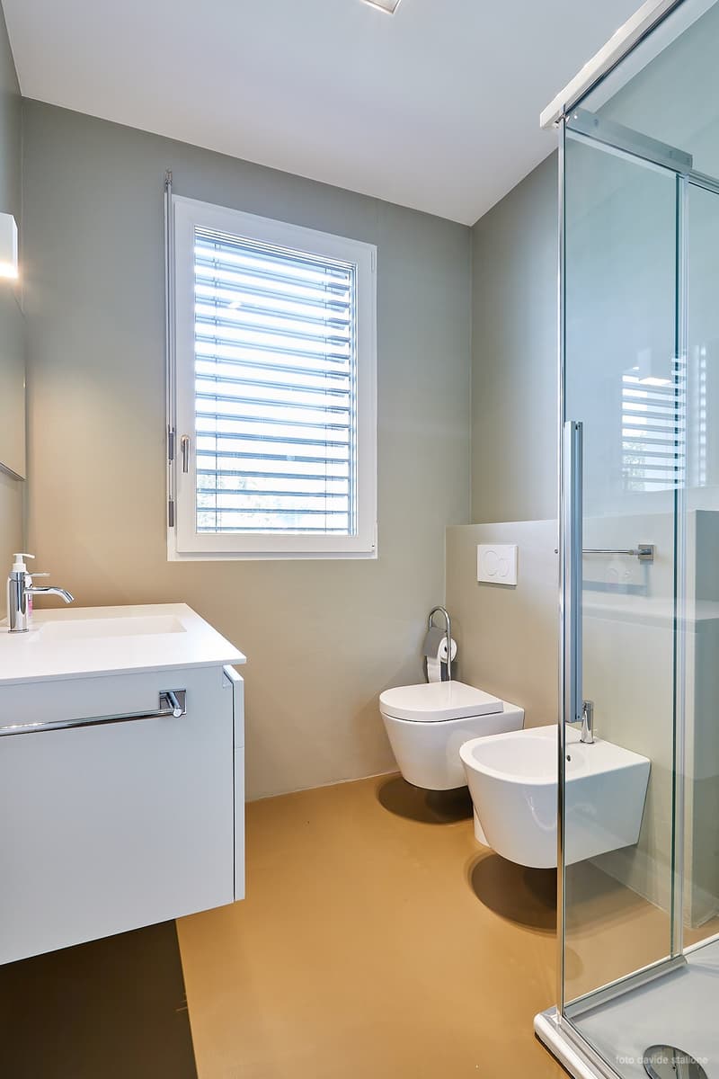 Modern bathroom with white vanity, toilet, bidet, and glass shower enclosure. Large window with horizontal blinds provides natural light.