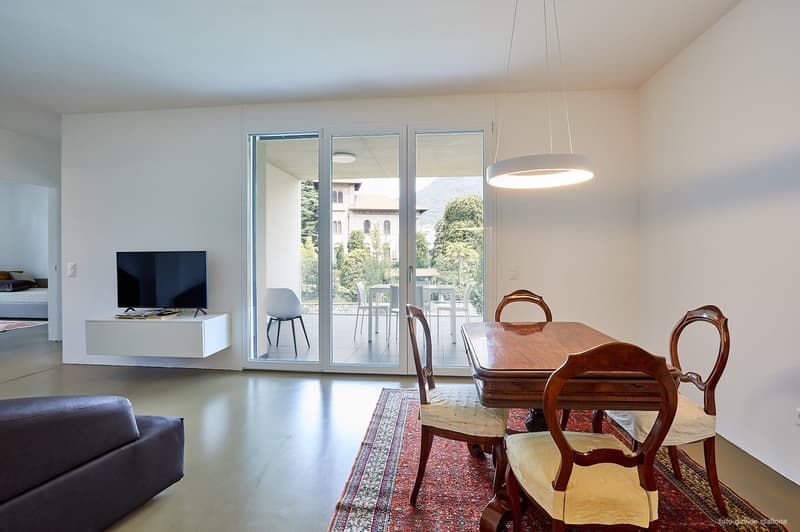 Spacious open-plan living and dining area with large windows providing a view of the outdoor area. The living room features a TV stand and a sofa, while the dining area has a wooden table with chairs. The room has a modern and minimalist design with a pen