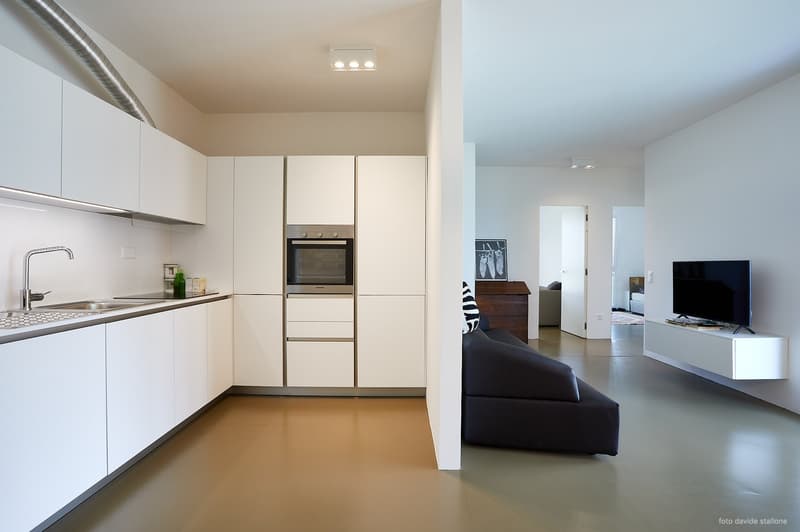 Modern, minimalist kitchen with white cabinets, stainless steel appliances including a dishwasher, and a sleek countertop. The kitchen opens up to a living room area with a black sofa and a TV stand.