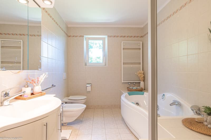 Spacious bathroom with beige tile walls, white vanity with sink, toilet, bathtub, and towel warmer. The room has a large window providing natural light.