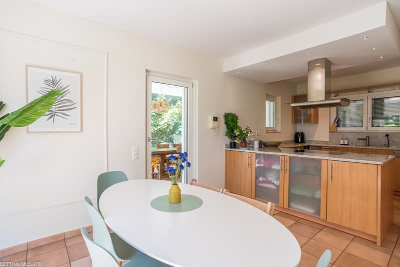 Open-concept kitchen and dining area with white walls, hardwood floors, and large windows overlooking a balcony or terrace. The kitchen features wooden cabinets, a granite countertop, and a built-in dishwasher. The dining table has a white top and mint gr