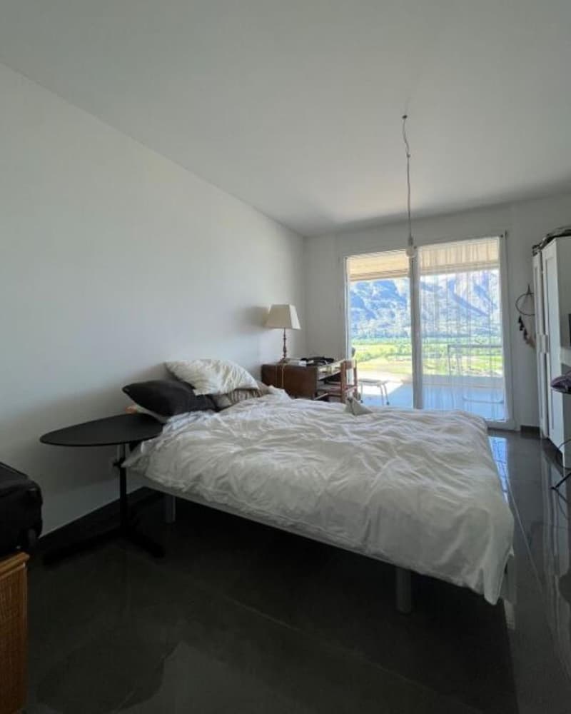 minimalist bedroom, gray floors, white walls, bed with white sheets, table with lamp, sliding glass doors leading to balcony with view
