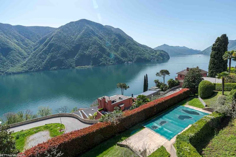 Luxury villa with a stunning view of the lake surrounded by mountains, with a swimming pool, balcony, and lush landscaping.
