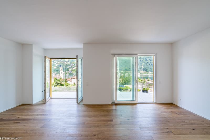 Spacious living room with large windows offering a scenic view of the surrounding landscape, including a church steeple in the distance. The room has hardwood floors and white walls, creating a bright and airy atmosphere.
