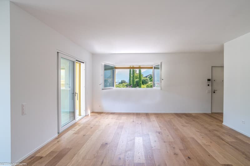 Spacious living room with large windows providing a view of the outdoor greenery, hardwood flooring, and a sliding glass door leading to a balcony or terrace.
