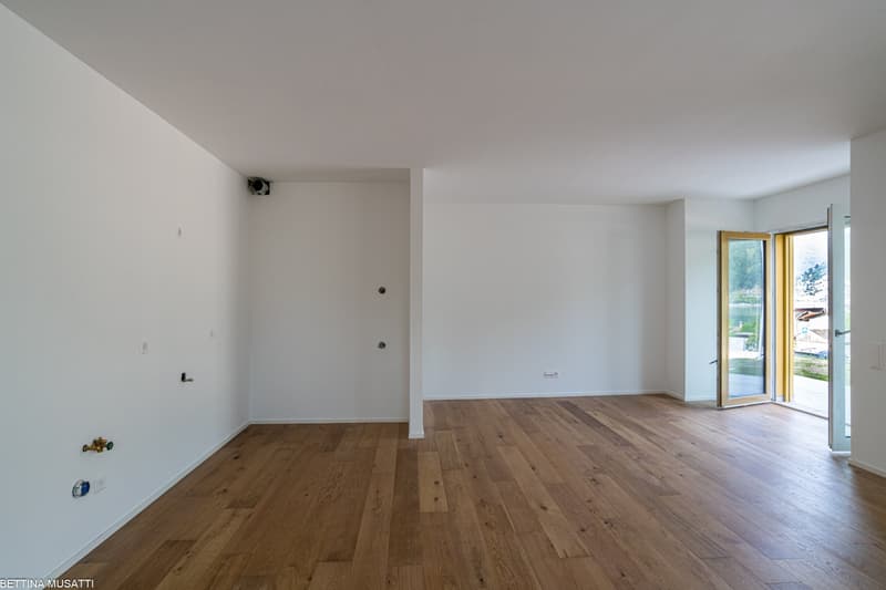 Spacious open-plan living room with hardwood flooring, white walls, and a large sliding glass door leading to a balcony or terrace with a view of the outdoors.