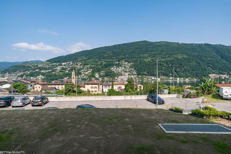 The image shows a scenic view of a town nestled in the mountains. The town has a mix of residential and commercial buildings, with a church steeple visible in the center. There is a parking lot in the foreground, and the surrounding hills are covered in l