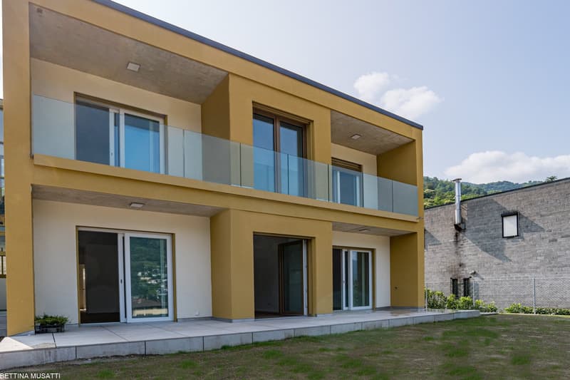 Two-story modern house with yellow exterior walls, large windows, and a balcony on the second floor. The house has a paved entrance and is surrounded by a grassy area.