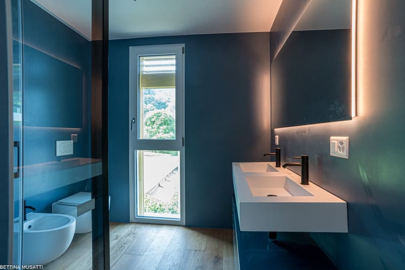 Modern bathroom with blue and dark gray walls, large window with a view, white sink vanity, and wooden flooring.