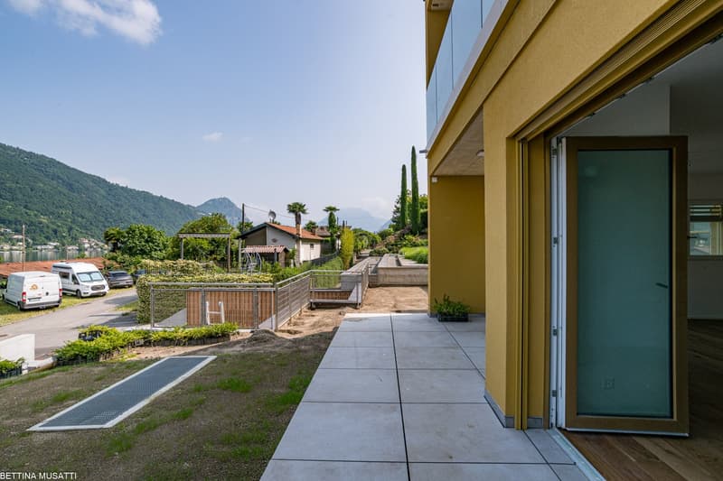 The image shows the exterior of a modern building with a balcony or terrace overlooking a scenic landscape. The building has a yellow facade and large windows. The surrounding area includes lush greenery, such as trees and shrubs, as well as a paved walkw