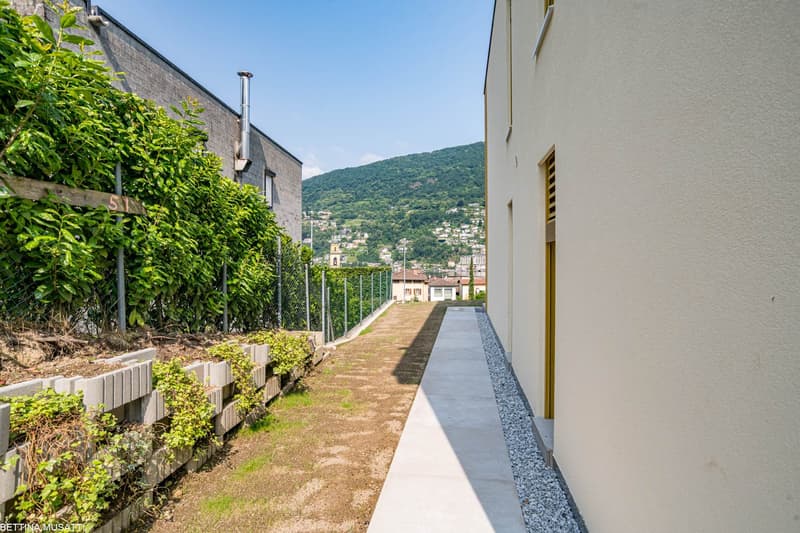 The image shows the exterior of a building with a walkway along the side. The walkway has a concrete path with gravel on the sides, and there are lush green plants and vegetation along the walls of the building. The building appears to have a balcony or t