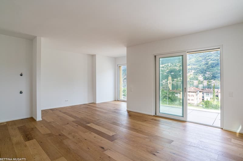 Spacious living room with large windows offering a scenic view of the surrounding landscape, including a church steeple and buildings. The room has hardwood flooring and white walls, creating a bright and airy atmosphere.