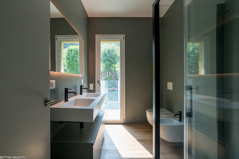 Modern bathroom with double vanity sink, black faucets, glass shower enclosure, and large windows overlooking a garden or outdoor area
