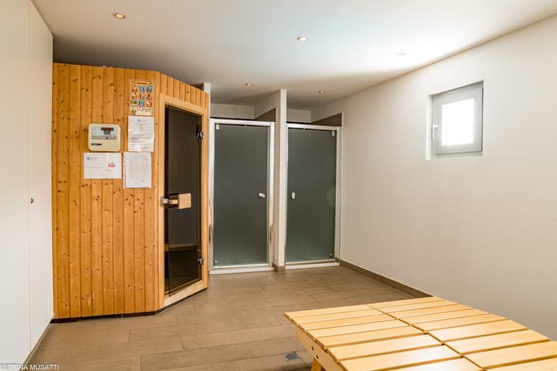 The image shows a sauna room with wooden paneling on the walls, a sauna unit, and two glass doors leading to what appears to be a changing area or shower. There is also a small window and a tiled floor in the room.