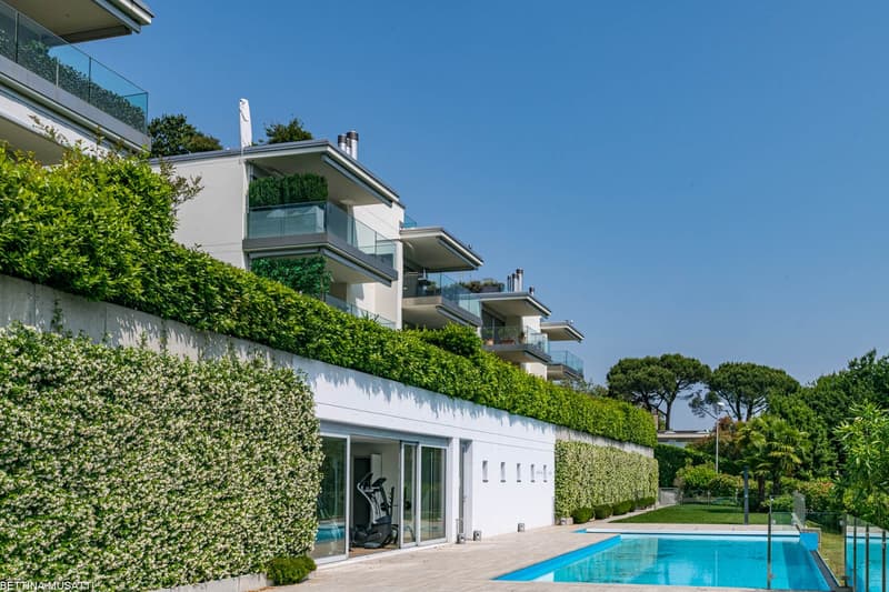 Multi-story apartment building with glass balconies, surrounded by lush greenery and landscaping. The building has a swimming pool visible in the foreground.