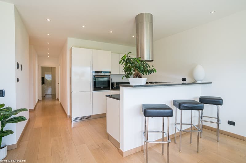 Spacious, modern kitchen with white cabinets, stainless steel appliances including a dishwasher, and a central island with bar stools. The kitchen opens up to a hallway and has hardwood floors throughout.