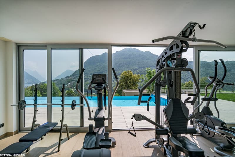 The image shows a well-equipped home gym with a variety of exercise equipment, including a treadmill, weight machines, and other fitness gear. The gym has large windows that provide a panoramic view of the surrounding mountains and a swimming pool outside