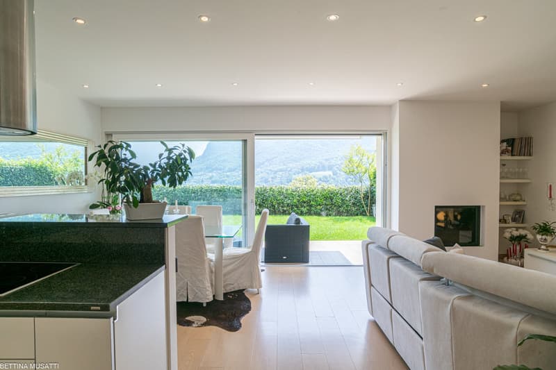 Spacious open-plan living area with large windows offering a scenic view of the surrounding greenery. The room features a modern kitchen with a glass-topped dining table, a comfortable sofa, and a potted plant adding a touch of nature. The floor is made o