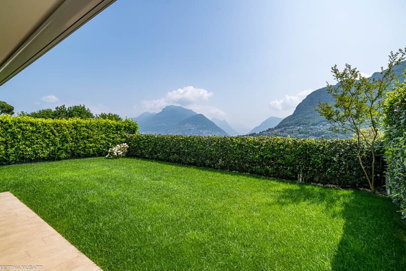 The image shows a well-manicured lawn surrounded by a neatly trimmed hedge. In the background, there are lush green mountains and a clear blue sky with some clouds. The scene appears to offer a beautiful view of the surrounding landscape.