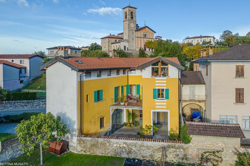 Two-story colorful house with yellow exterior, green shutters, and a balcony. The house is surrounded by other buildings and has a church tower visible in the background.